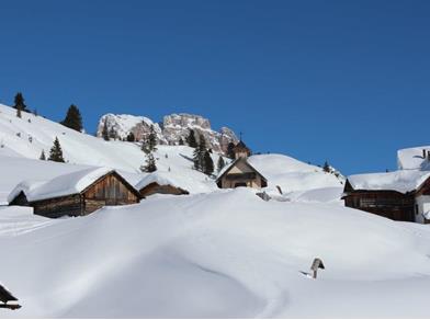 Snow-covered alpine village with wooden huts and chapel, surrounded by mountains under clear skies.